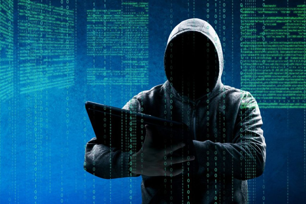 Hooded anonymous hacker figure using a laptop against a dark digital code background